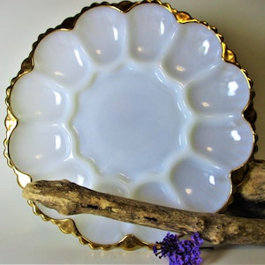 May include: A white, scalloped serving dish with a gold rim. The dish has a central circular area surrounded by 12 individual compartments. A piece of driftwood and purple flowers are in the foreground.