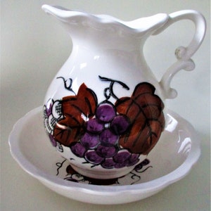 May include: White ceramic pitcher and matching bowl with a grapevine design. The pitcher has a brown and purple grape design with green leaves. The bowl has a smaller grape design.