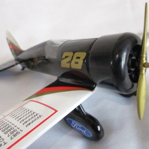 May include: Black and white toy airplane with a gold propeller and the number "28" on the side. The airplane has a red and gold stripe on the wing and a blue "Ford" logo on the bottom.