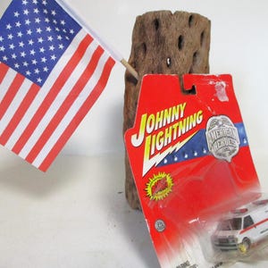 May include: A red, white, and blue American flag is displayed next to a toy ambulance in a plastic package. The package is red and white with the words "Johnny Lightning" and "American Heroes" printed on it.