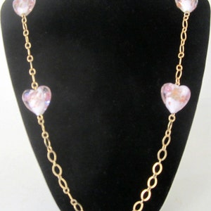 May include: A gold chain necklace with four pink heart-shaped glass beads. The beads have a white and pink floral pattern.