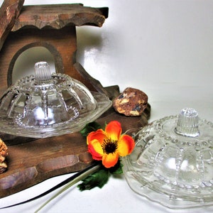 May include: Two clear glass butter dishes with lids, featuring a decorative pattern. The dishes sit on a wooden tray with a carved design, accompanied by a colorful flower and small rocks. A vintage kitchenware item.