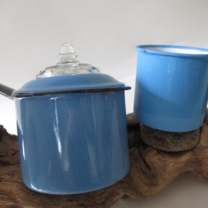 May include: A blue enamelware saucepan with a glass lid and a blue enamelware mug. Both are sitting on a piece of driftwood.
