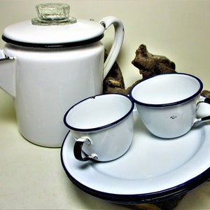 May include: A white enamel coffee pot with a black rim and a glass lid, two white enamel mugs with black rims, and two white enamel saucers with black rims.