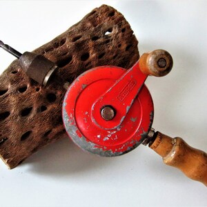 May include: A red hand crank drill with a wooden handle and a black drill bit. The drill is resting on a piece of wood with holes in it.