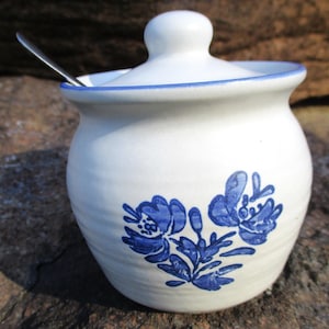 May include: White ceramic sugar bowl with a blue floral design and a lid. The bowl has a spoon resting on the rim.