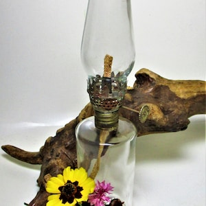 May include: A clear glass oil lamp with a metal wick holder and a glass reservoir. The lamp is sitting on a piece of driftwood with a few yellow, pink, and blue flowers.