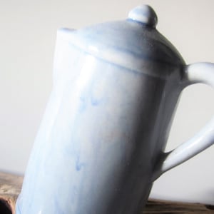 May include: A light blue ceramic pitcher with a lid and handle. The pitcher has a rounded body and a curved handle. The lid has a small knob on top. The pitcher is glazed with a marbled effect.