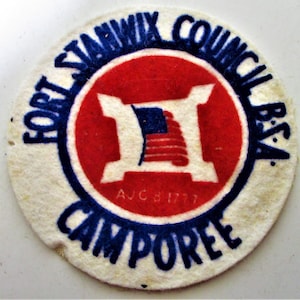 May include: A white felt patch with blue text that reads "Fort Stanwix Council BSA Campore" around a red circle with a white "H" inside. The "H" has a small American flag inside. The text "A.J.C. 8 1777" is printed in white inside the red circle.
