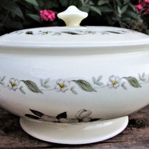 May include: Cream-colored ceramic serving dish with lid. The dish features a floral design with white flowers and green leaves. It has two handles and a knob on the lid. A classic piece for serving soups, stews, or other dishes.
