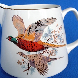 May include: White ceramic pitcher with a gold rim and a colorful illustration of two pheasants in flight. The pheasants are perched on a branch with green leaves and berries.