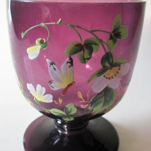 May include: A purple glass goblet with a hand-painted design of butterflies and flowers. The goblet has a wide bowl and a short stem.