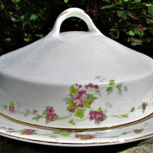 May include: A white ceramic butter dish with a floral design. The dish has a lid with a handle and a base with a gold trim.