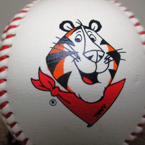 May include: White baseball with red stitching and a cartoon orange and black tiger wearing a red bandana with the word "Tony" on it.