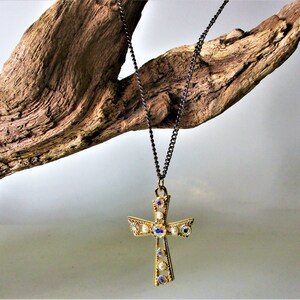 May include: A gold cross pendant necklace with iridescent rhinestones and a black chain. The necklace is hanging from a piece of driftwood.