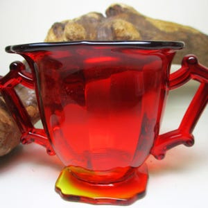 May include: A red glass sugar bowl with two handles. The bowl has a fluted design and a yellow base.