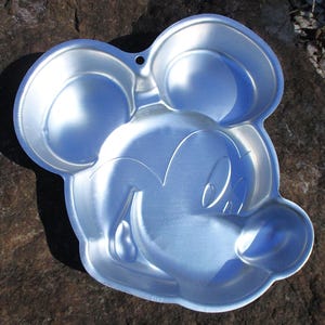 May include: A silver-colored, Mickey Mouse-shaped cake pan. The pan features the iconic mouse's face, including large ears, eyes, nose, and a smiling mouth. The pan has a small hole for hanging and is made of a shiny, reflective metal.