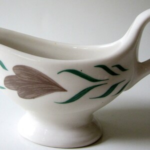 May include: White ceramic gravy boat with a green and brown floral design. The boat has a handle and a base.