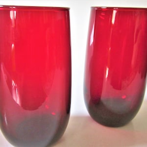 May include: Two red-tinted, stemless drinking glasses. The glasses have a rounded, cylindrical shape with a darker base. The vibrant red color transitions to a deeper shade at the bottom. These glasses are suitable for water, juice, or other beverages.