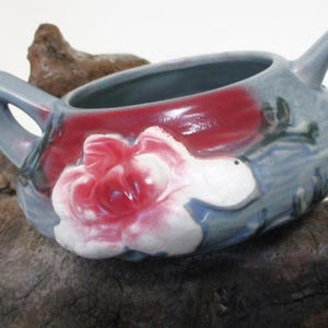 May include: A small, blue and red ceramic teapot with a floral design. The teapot has two handles and a white flower with pink accents.