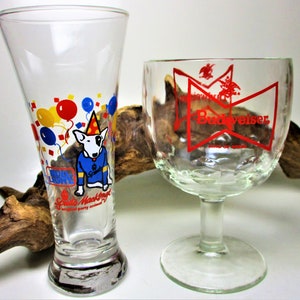 May include: Two glass beer mugs. The mug on the left is clear with a cartoon dog wearing a party hat and surrounded by balloons. The mug on the right is clear with a red Budweiser logo.