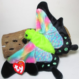 May include: A green and black butterfly Beanie Baby with rainbow wings and a Ty tag. The butterfly is sitting on a piece of driftwood.