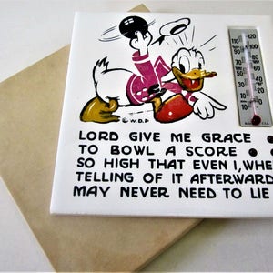 May include: A white ceramic tile with a cartoon image of a duck bowling. The duck is wearing a pink shirt and a red hat. The tile has a thermometer on the right side and a quote that reads: "LORD GIVE ME GRACE TO BOWL A SCORE. SO HIGH THAT EVEN I, WHEN TELLING OF IT AFTERWARDS MAY NEVER NEED TO LIE."