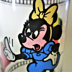 May include: A cartoon image of Minnie Mouse wearing a blue dress and a yellow bow. She has a pink nose and is holding up two fingers. The image is on a glass.