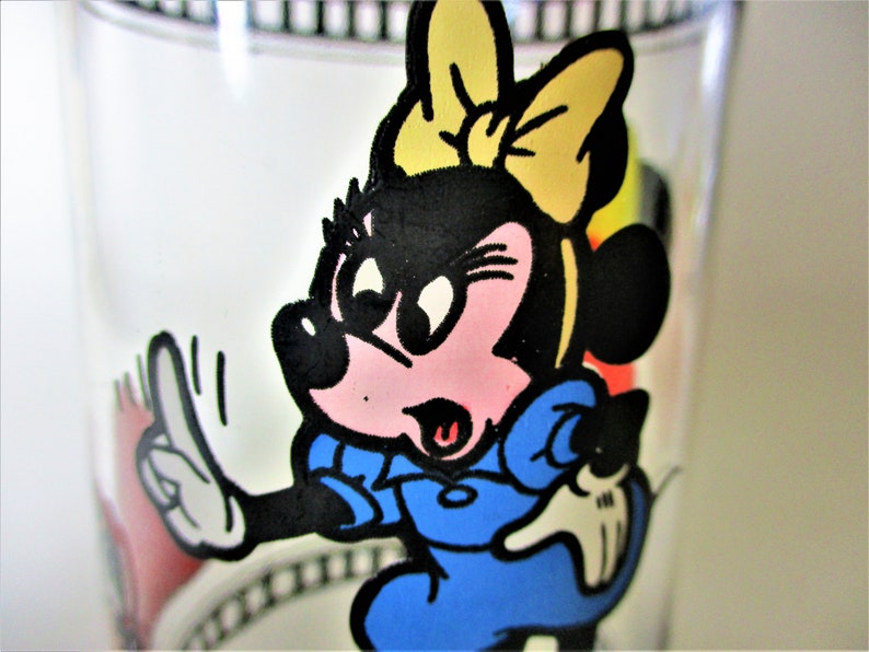 May include: A cartoon image of Minnie Mouse wearing a blue dress and a yellow bow. She has a pink nose and is holding up two fingers. The image is on a glass.
