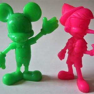 May include: Green plastic figurine of Mickey Mouse with a raised hand, standing next to a pink plastic figurine of Pinocchio with a raised thumb.