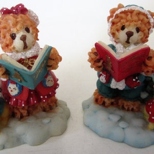 May include: Two ceramic figurines of teddy bears dressed in Christmas attire, each holding a book and standing on a snowy base. One bear is wearing a red and white dress and the other is wearing a green and white outfit. Both bears have a sweet expression on their faces.