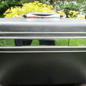 May include: A black hardshell travel case with a textured exterior finish. The case features a silver-toned metallic band running horizontally across the center, which includes two integrated key locks and a central reflective chrome-like panel.