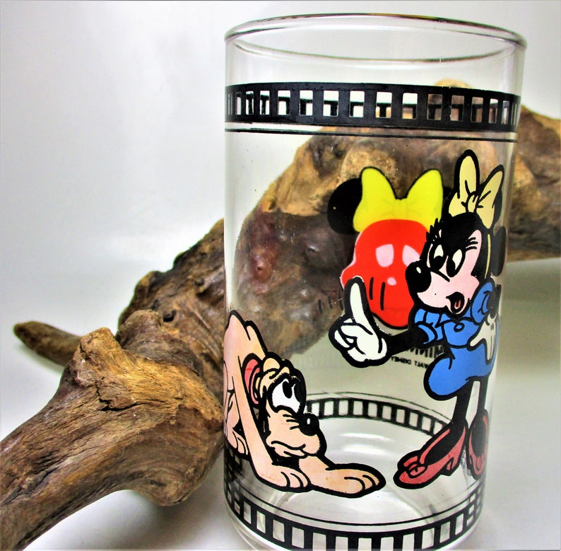 May include: A clear glass with a black and white film strip design around the top. The glass features a cartoon image of Minnie Mouse and Pluto. Minnie Mouse is wearing a red dress with yellow polka dots and a yellow bow. Pluto is brown and white and is lying down. The text "Disneyland" is visible on the glass.