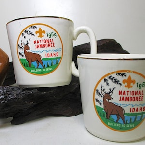 May include: Two white ceramic mugs with a gold trim. Both mugs feature a colorful graphic of a deer and text that reads "1969 National Jamboree Idaho Building to Serve".