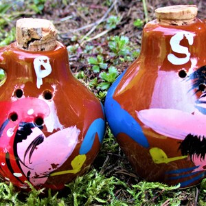 May include: A set of two brown ceramic salt and pepper shakers with cork tops. The shakers are painted with colorful designs of people with pink faces and black hair. The salt shaker has a white "P" painted on it and the pepper shaker has a white "S" painted on it.