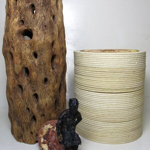 May include: A black figurine of a person sitting on a small, red, rocky surface. The figurine is in front of a large, brown, cylindrical piece of wood with holes and a stack of white, round coasters.