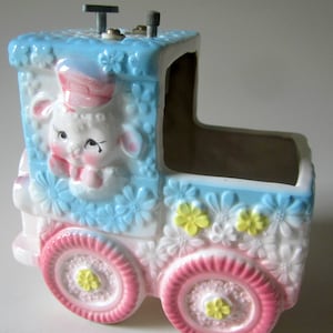 May include: A ceramic musical train with a lamb face on the front. The train is decorated with pink, blue, and white flowers. The train has a winding key on the top.