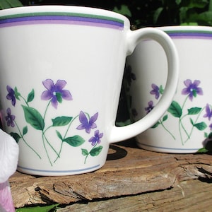 May include: Two white ceramic mugs with a floral design featuring purple flowers and green leaves. The mugs have a handle and a decorative band of purple, green, and blue near the rim. The mugs are on a wooden surface.