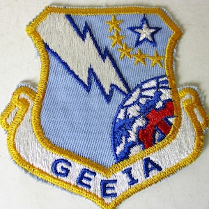 May include: Embroidered patch with a light blue shield design, featuring a white lightning bolt, gold stars, and a blue and red globe. The patch has a gold border and the letters "GEEIA" in white.