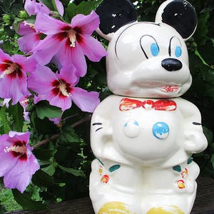 May include: A white ceramic Mickey Mouse cookie jar with black ears, blue eyes, and a red bow tie. The jar is decorated with colorful accents and sits in front of pink flowers.