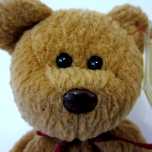 May include: A brown teddy bear with black eyes and a brown nose. The bear is wearing a red ribbon around its neck. The bear has a Ty tag attached to its ear.