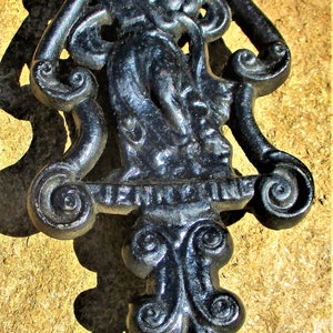 May include: Black cast iron door knocker with a decorative design featuring a figure and scrollwork. The word "JENNINGS" is visible on the knocker. The knocker is mounted on a textured, tan surface.