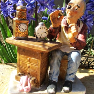 May include: A figurine of a clockmaker at work, seated at a wooden workbench. A gold-colored clock and a small silver clock sit on the table. The clockmaker wears a red shirt, blue jeans, and a white apron. Purple flowers are in the background.