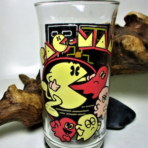 May include: A clear glass with a Pac-Man design. The glass features Pac-Man, ghosts, and the words "Pac-Man" in yellow, red, and black. The glass is on a wooden surface.