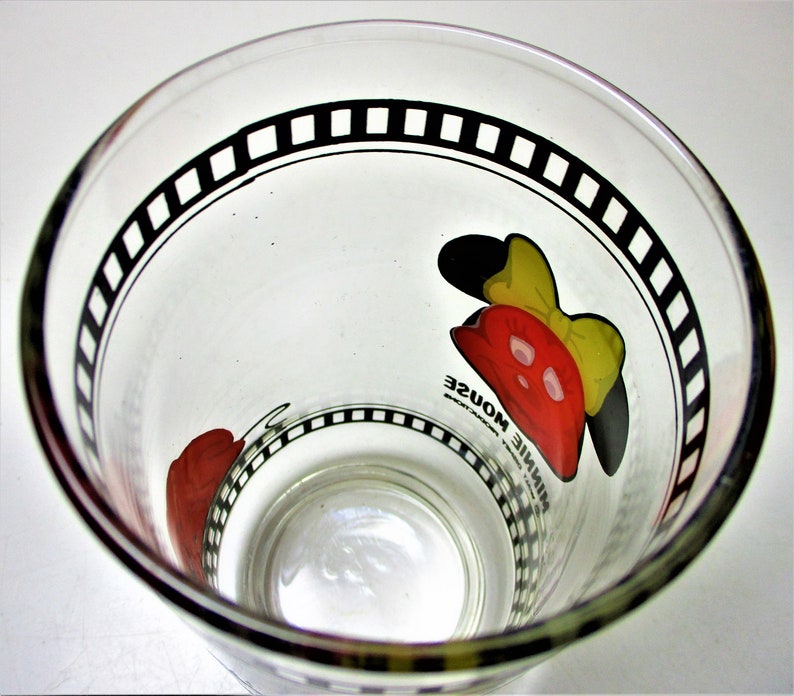 May include: A clear glass drinking glass with a black and white film strip design around the rim. The glass features a red and yellow cartoon image of Minnie Mouse with the text "Minnie Mouse" printed on the glass.