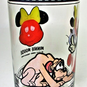 May include: A clear glass drinking glass with a black and white film strip design around the top and bottom. The glass features cartoon images of Mickey Mouse, Minnie Mouse, and Pluto. The text "Minnie Mouse" is printed on the glass.