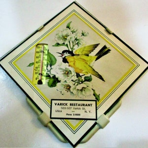 May include: A vintage thermometer with a yellow bird and white flowers on a white background. The thermometer is in a diamond-shaped frame with a green border. The frame has a white card with the text "VARICK RESTAURANT 503-507 Varick St. Utica N.Y. Phone 2-9569".