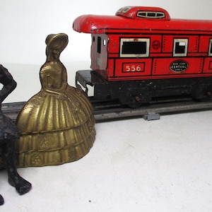 May include: A red toy train car with black wheels and a black undercarriage. The car has a white number "556" on each side and the words "New York Central Lines" in white on the side. The train car is on a gray track with a small, gold-colored figurine of a woman in a dress.