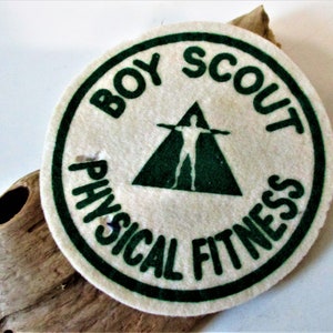 May include: A white felt patch with a green border. The patch has a green triangle with a white silhouette of a person with arms outstretched. The text "BOY SCOUT PHYSICAL FITNESS" is written in a green circle around the triangle.