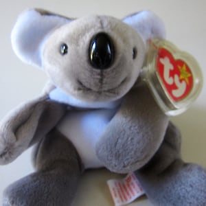 May include: A gray and blue plush koala bear with a Ty Beanie Baby tag attached to its ear. The tag is red with a white star and the word "Ty" in white.